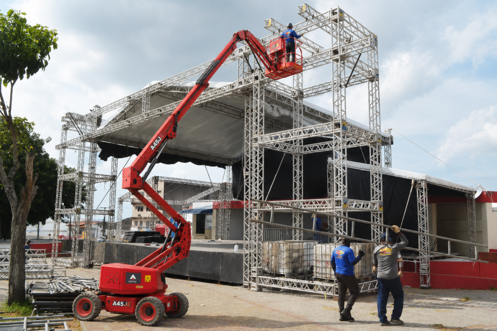 Operador em plataforma articulada monta estrutura metálica de palco enquanto equipe trabalha na montagem do evento.