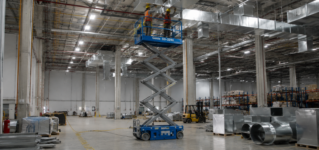 Plataforma tesoura elétrica eleva dois trabalhadores para instalação de dutos em galpão industrial.