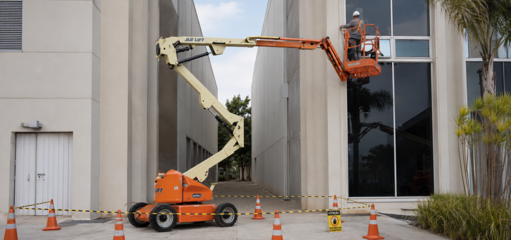 Plataforma articulada realiza manutenção na fachada de edifício comercial com área isolada por cones de segurança.