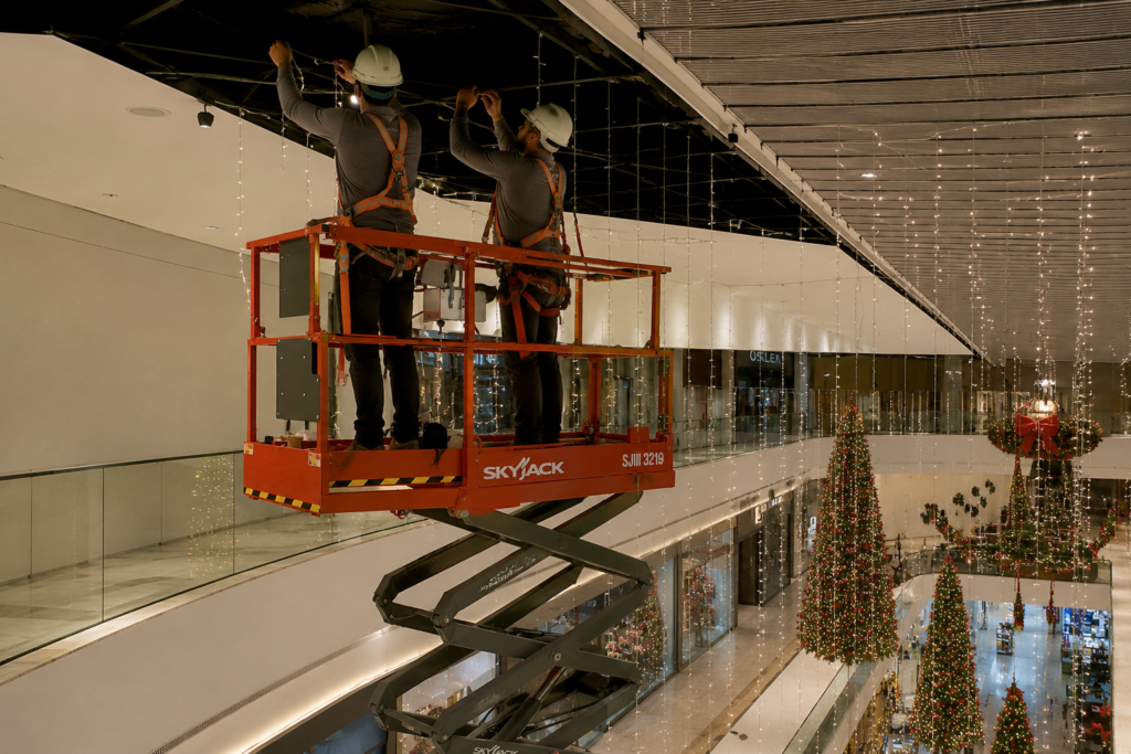 Técnicos com EPI utilizam plataforma tesoura para instalar iluminação natalina no teto de um shopping.