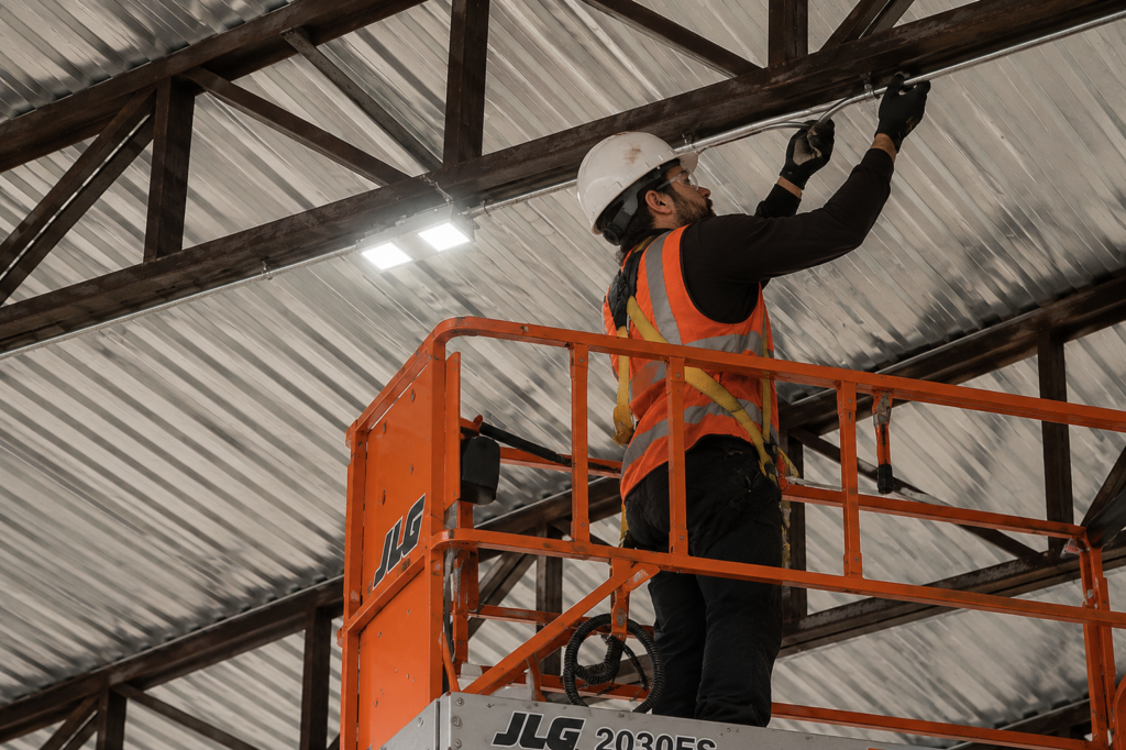 Técnico com EPI realiza instalação elétrica em altura utilizando plataforma elevatória tipo tesoura.
